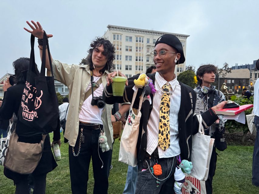 Two people stand outdoors in a park, smiling and holding tote bags and drinks, with other people and buildings visible in the background.