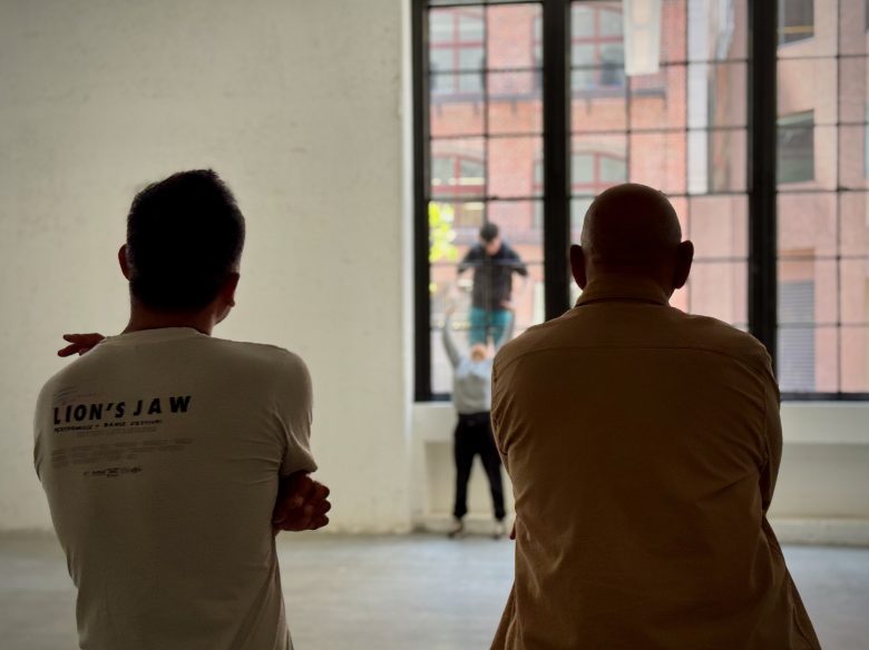 Two people stand in the foreground facing large windows, while two others work outside on the windows, with a brick building visible through the glass.