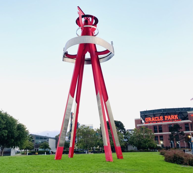Tall red and white abstract sculpture on a grassy area near Oracle Park, with trees and buildings in the background under a clear sky.