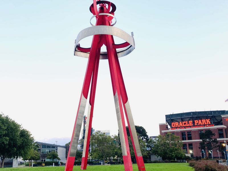 Tall red and white abstract sculpture on a grassy area near Oracle Park, with trees and buildings in the background under a clear sky.