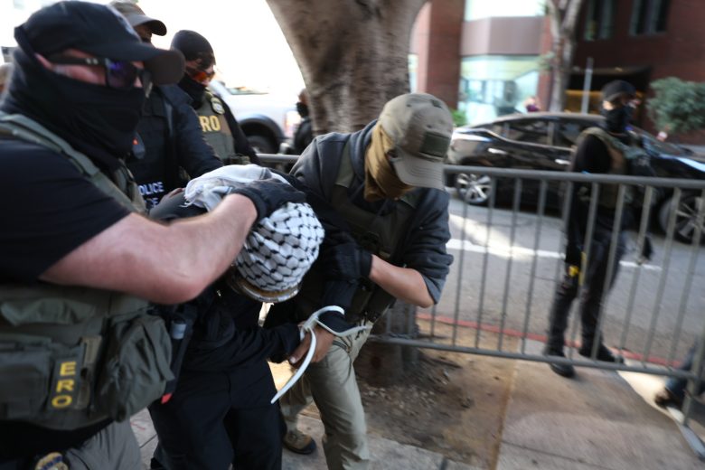 Several masked law enforcement officers detain and handcuff a person in front of a metal barricade on a city sidewalk.