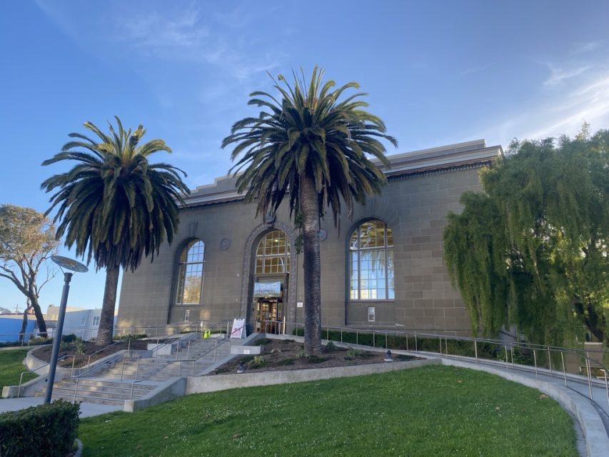 A large building with tall arched windows is flanked by two palm trees, with a ramp and stairs leading to the entrance. The sky is clear and blue.
