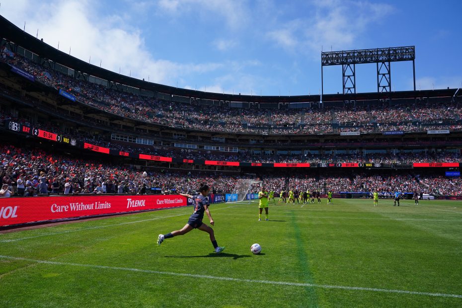 A soccer player kicks a ball from the sideline during a match in a large stadium filled with spectators on a sunny day.