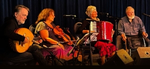 Four musicians sit on stage with instruments, including a guitar, violin, accordion, and a percussion instrument, performing in front of dark blue curtains.