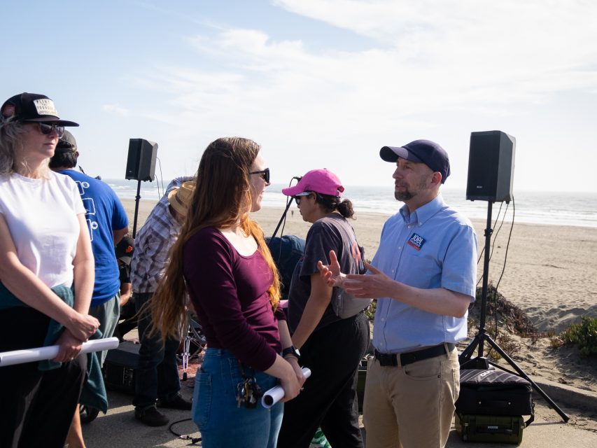A group of people stand and talk near speakers and equipment on a beach, with the ocean and sky visible in the background.