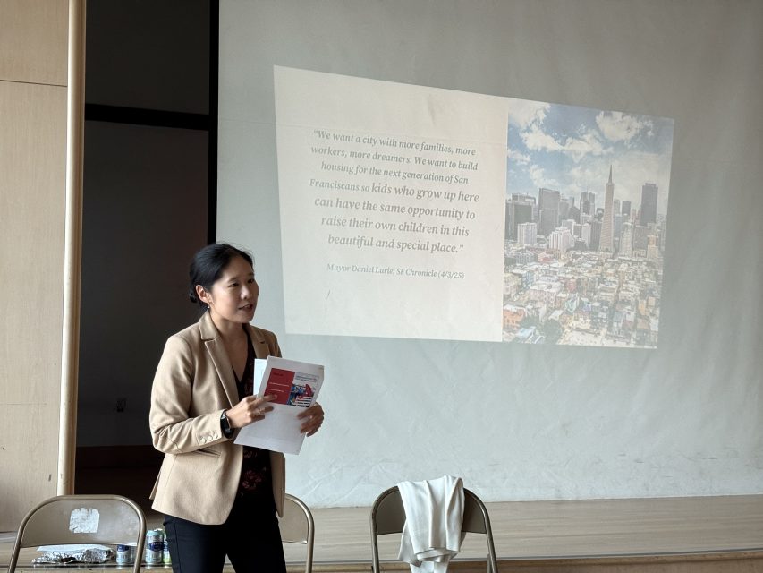 A woman stands in front of a projected slide with a quote about San Francisco, holding papers and speaking to an audience in a classroom or meeting space.