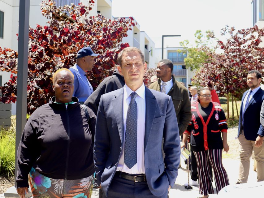 A group of people, including a man in a suit standing in front, gather outdoors near buildings and trees on a sunny day.