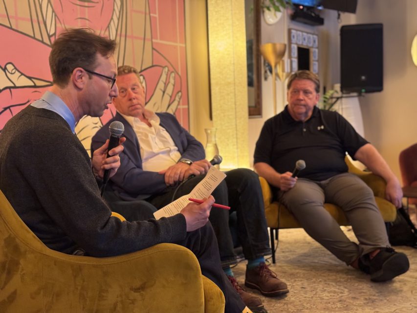 Three men seated indoors; one holds a microphone and reads from a paper, while the other two listen, each holding a microphone. A mural and speaker are visible in the background.