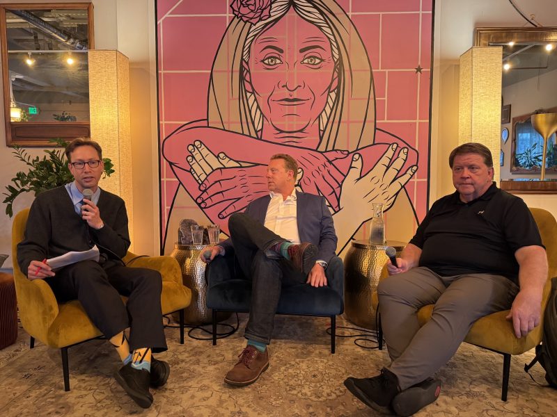 Three men sit in armchairs in front of a large pink mural of a woman with folded arms; one holds a microphone, appearing to moderate a discussion.