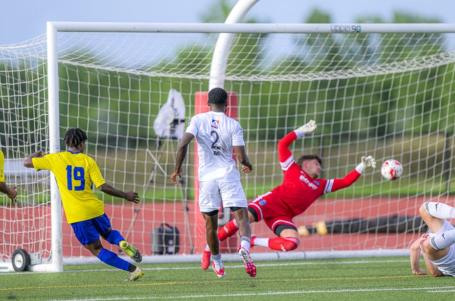 A soccer player in a yellow jersey shoots at the goal as a goalkeeper in red dives to block, with defenders in white nearby and the ball heading towards the net.