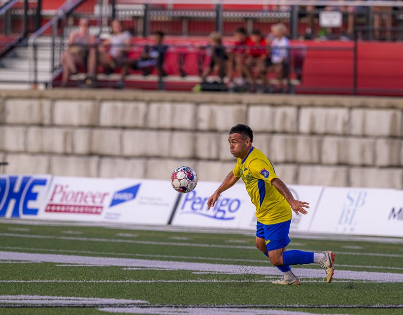 A soccer player in a yellow jersey and blue shorts runs toward a ball on a field, with blurred spectators seated in the background.
