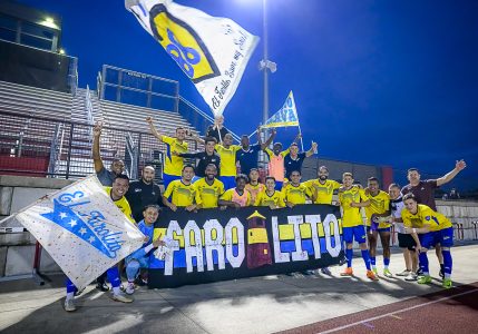 A soccer team in yellow uniforms poses together at night in front of empty stadium stands, holding flags and a banner reading "Faro Leito.