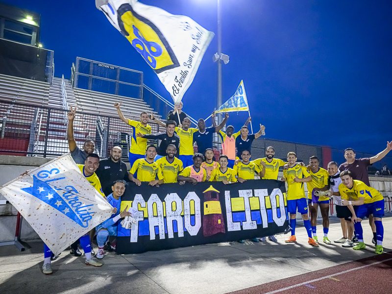 A soccer team in yellow uniforms poses together at night in front of empty stadium stands, holding flags and a banner reading "Faro Leito.