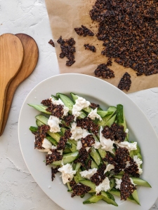 A white plate with sliced cucumbers, mozzarella cheese, and crumbled dark crisp topping sits next to wooden utensils and a tray with more dark crisp pieces on parchment paper.