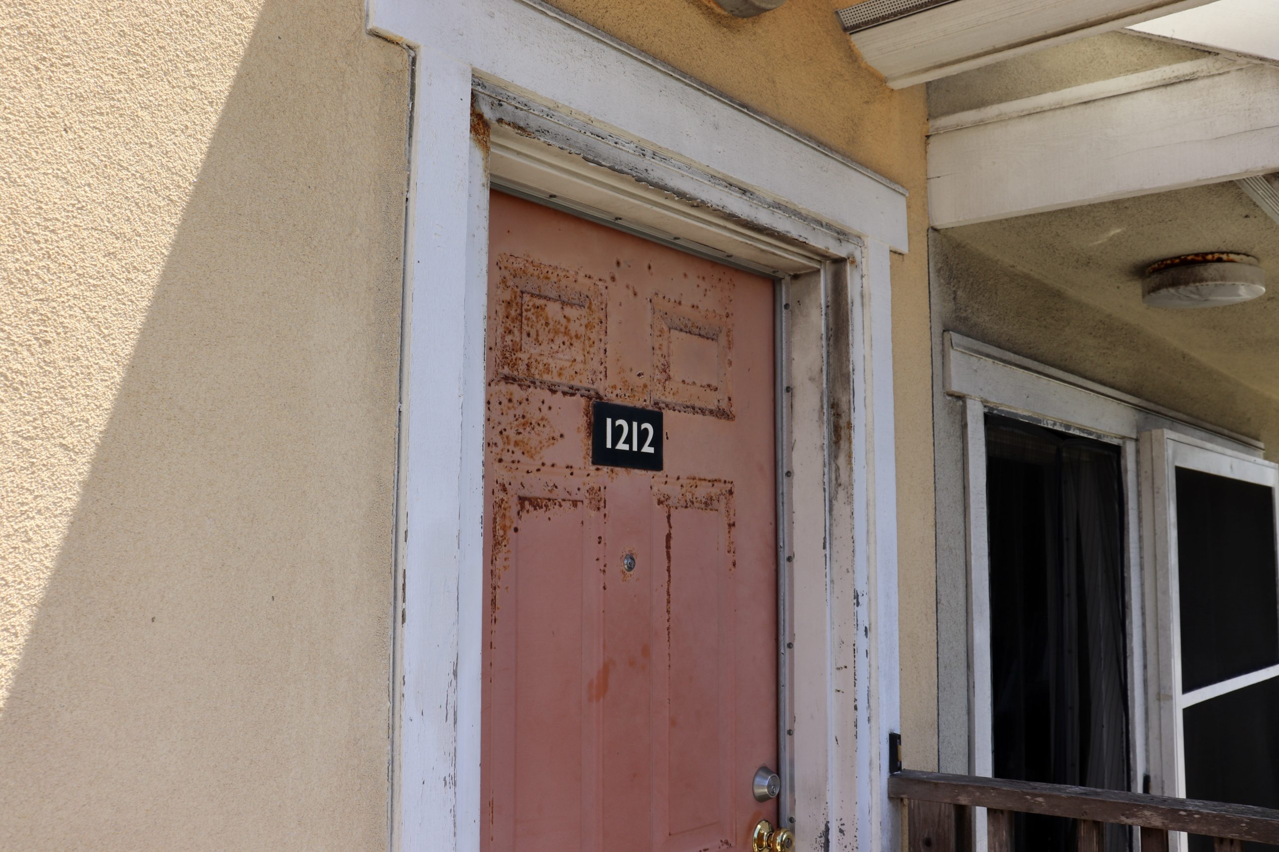 A weathered pink door with the number 1212 mounted above the center, surrounded by a white frame on a beige building.