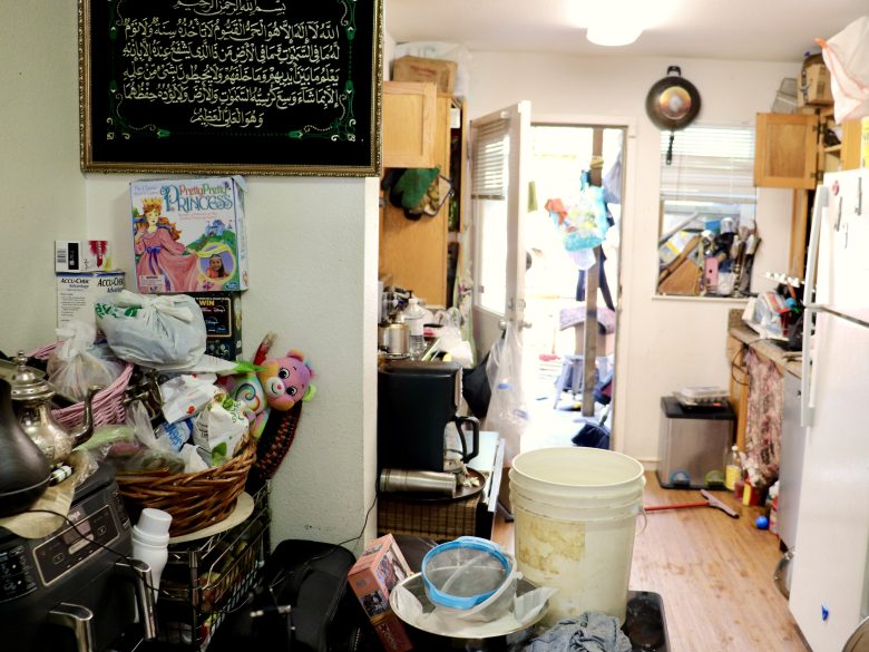 A cluttered kitchen with various household items, toys, and containers scattered on counters; a door at the back is open to a messy outdoor area.