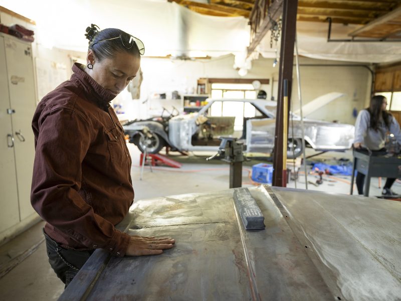 A person inspects and sands the surface of a car hood in an auto repair workshop, with another person working in the background.