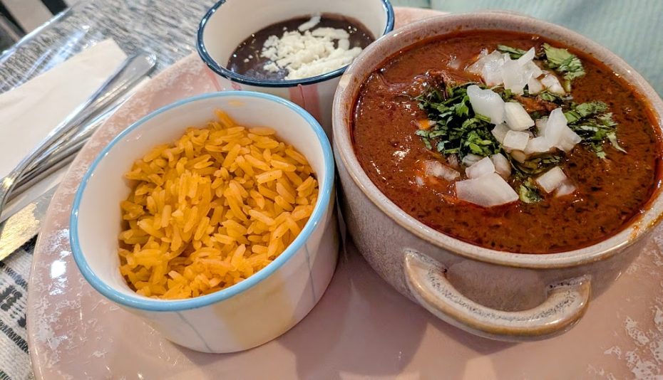 A bowl of red soup topped with chopped onion and cilantro, served with a side of Mexican rice and black beans garnished with cheese.