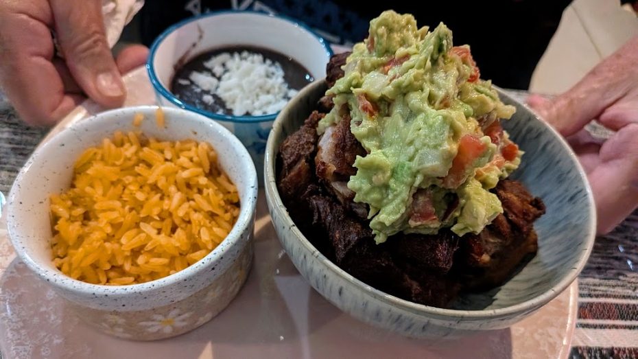 A bowl of carnitas topped with guacamole, served with sides of Mexican rice and black beans with cheese. Hands are holding the main bowl and a napkin.