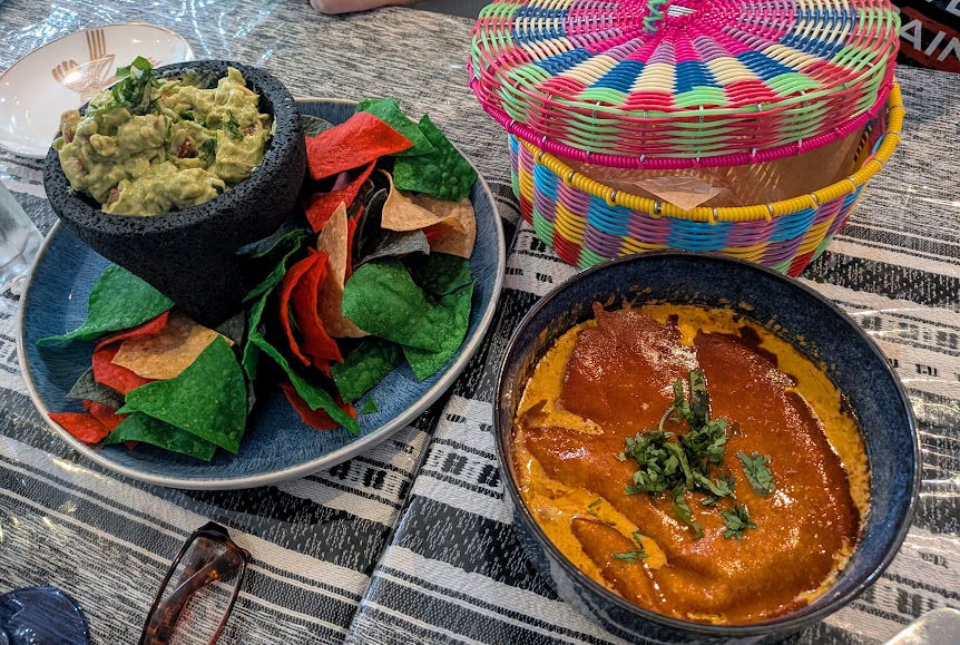 Tri-color tortilla chips with guacamole on a plate, a bowl of red sauce garnished with herbs, and a colorful woven basket on a striped tablecloth.