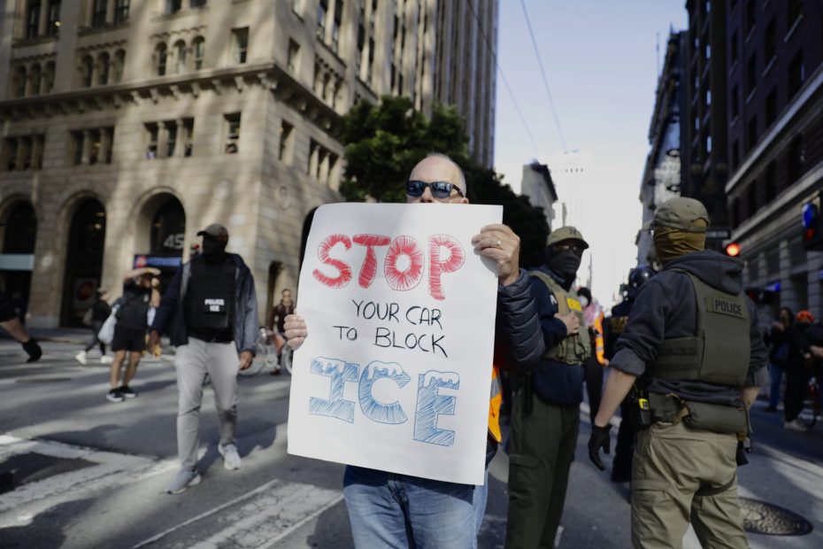 A person in sunglasses holds a sign reading "STOP YOUR CAR TO BLOCK ICE" during a street protest; other people and buildings are visible in the background.