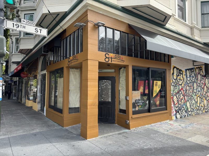 A corner restaurant with covered windows and a sign that reads "Grill" on 19th Street. The building has tan trim, a black door, and graffiti on the adjacent wall.