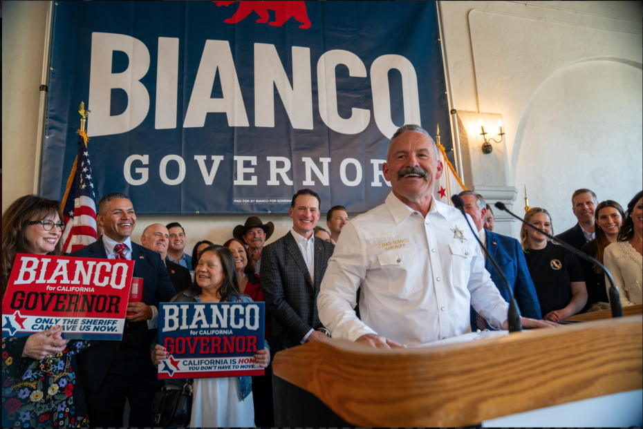 A man speaks at a podium in front of a large "Bianco Governor" banner, joined by people holding campaign signs and smiling, while the local Sheriff stands proudly among the supporters.