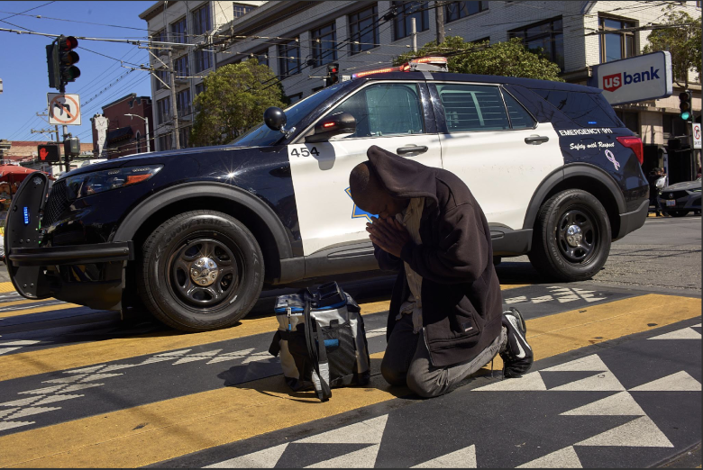 A person kneels on a crosswalk with hands pressed together in front of a parked police SUV on a city street, possibly in response to a recent shooting.