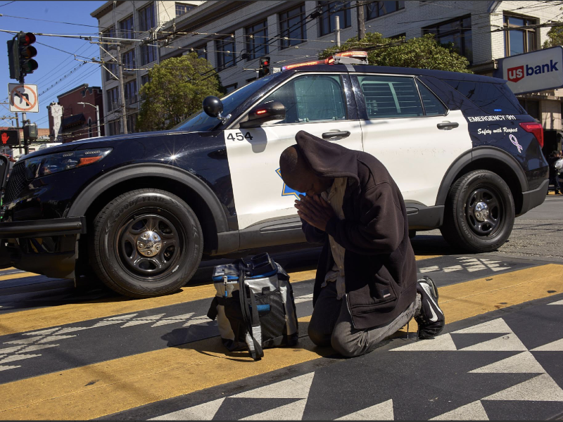 A person kneels on a crosswalk with hands pressed together in front of a parked police SUV on a city street, possibly in response to a recent shooting.