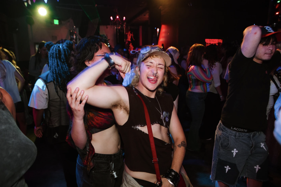 Person in a cut-off shirt flexes their arm and sticks out their tongue in a crowded, colorful club or party setting. Other people are dancing and mingling in the background.