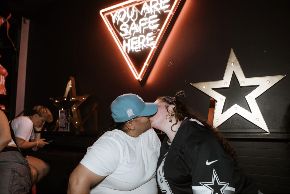 Two people kiss in front of a neon sign that reads "YOU ARE SAFE HERE," with star decorations on the wall and another person sitting in the background.