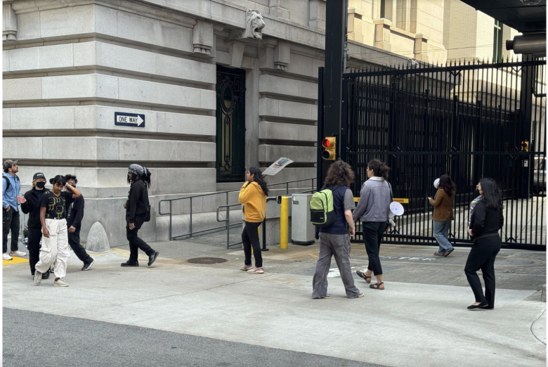 A group of people walk on a city sidewalk near a gated building, some holding megaphones and signs. A "One Way" sign and a traffic light are visible.