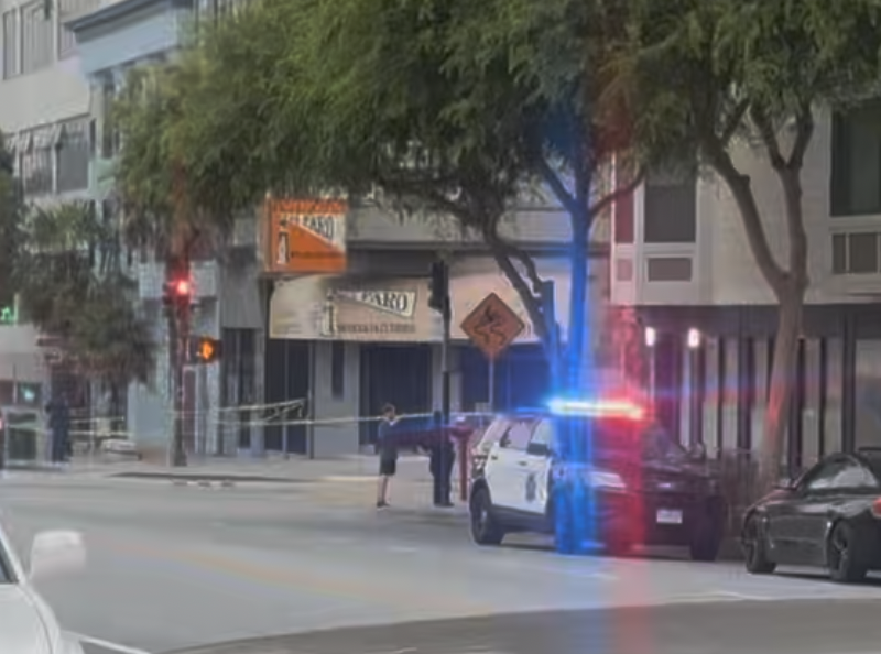 A police car with flashing lights is parked on a city street, yellow caution tape blocks the sidewalk, and a person stands nearby.