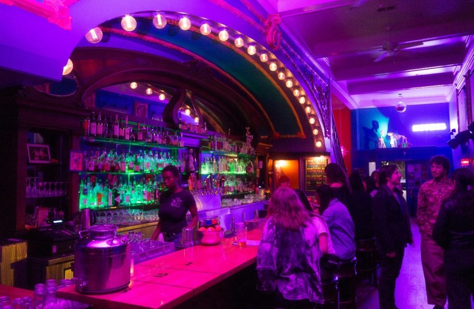 A dimly lit bar with colorful neon lights, bottles on shelves behind the counter, a bartender serving drinks, and patrons standing and sitting at the bar.