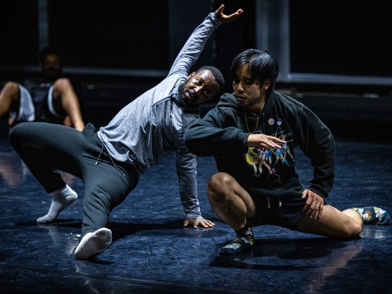 Two dancers perform on stage, one stretching an arm upward while leaning back, the other crouched with a bent arm, both focused and expressive in their movements.