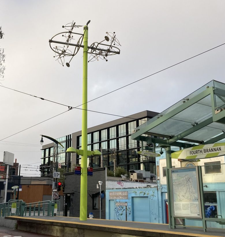 A green metal pole with an abstract sculpture on top stands at the Fourth/Brannan light rail station in an urban area with modern buildings in the background.