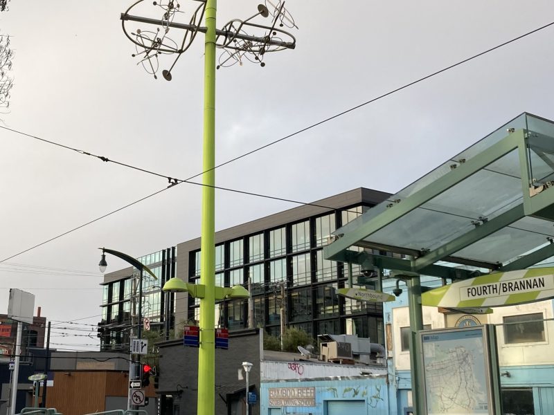 A green metal pole with an abstract sculpture on top stands at the Fourth/Brannan light rail station in an urban area with modern buildings in the background.