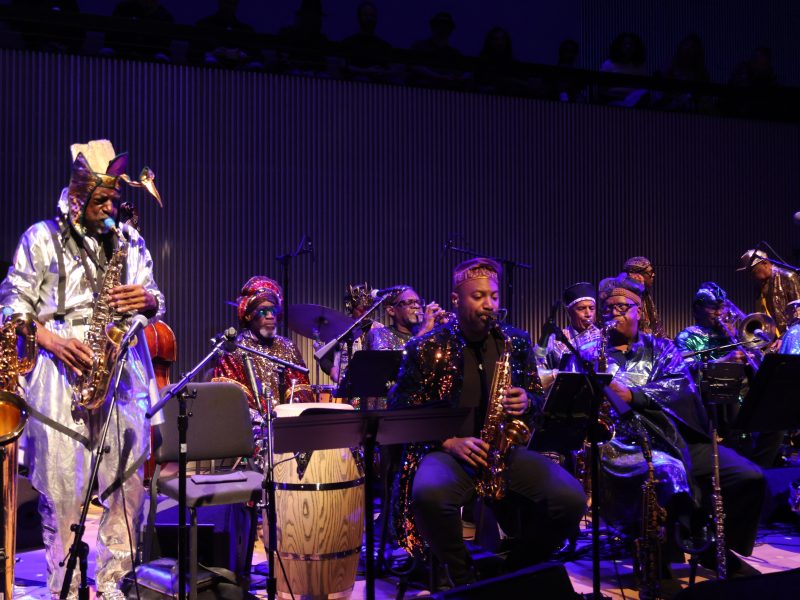 A large jazz band dressed in shiny, metallic costumes performs on stage with saxophones, drums, and other instruments.