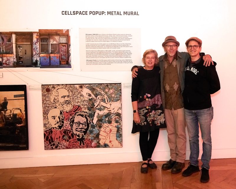 Three people stand together in front of a gallery wall displaying artwork and a sign reading "Cellspace Popup: Metal Mural.