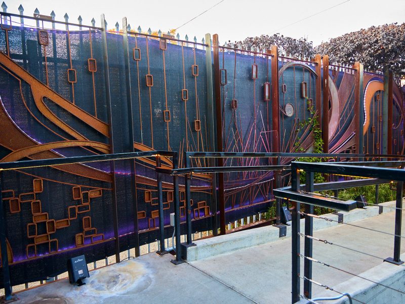 A decorative metal fence with abstract geometric and flowing designs, illuminated by purple lights, behind a concrete sidewalk and metal railing.