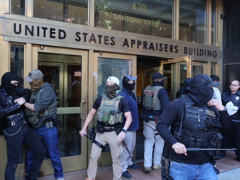 Law enforcement officers detain and search individuals outside the United States Appraisers Building, with police presence and activity near the entrance.