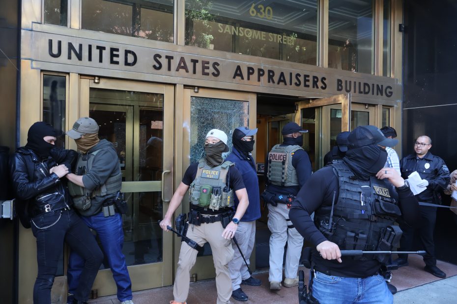Law enforcement officers detain and search individuals outside the United States Appraisers Building, with police presence and activity near the entrance.