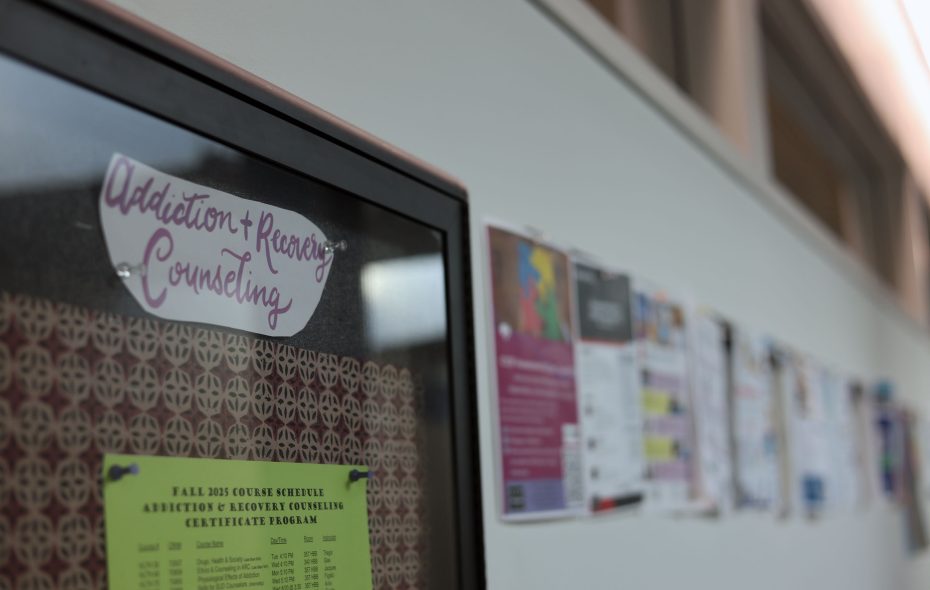 A bulletin board displays a sign reading "Addiction + Recovery Counseling" along with various flyers and notices pinned to the wall in a hallway.