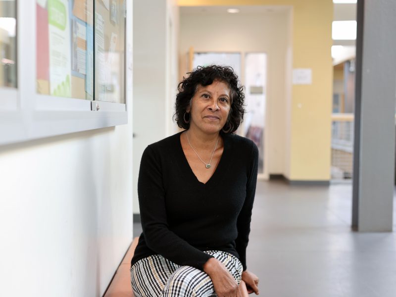 A woman with curly hair and a black sweater sits on a bench in a hallway, looking slightly upward. Bulletin boards and papers are visible on the wall beside her.