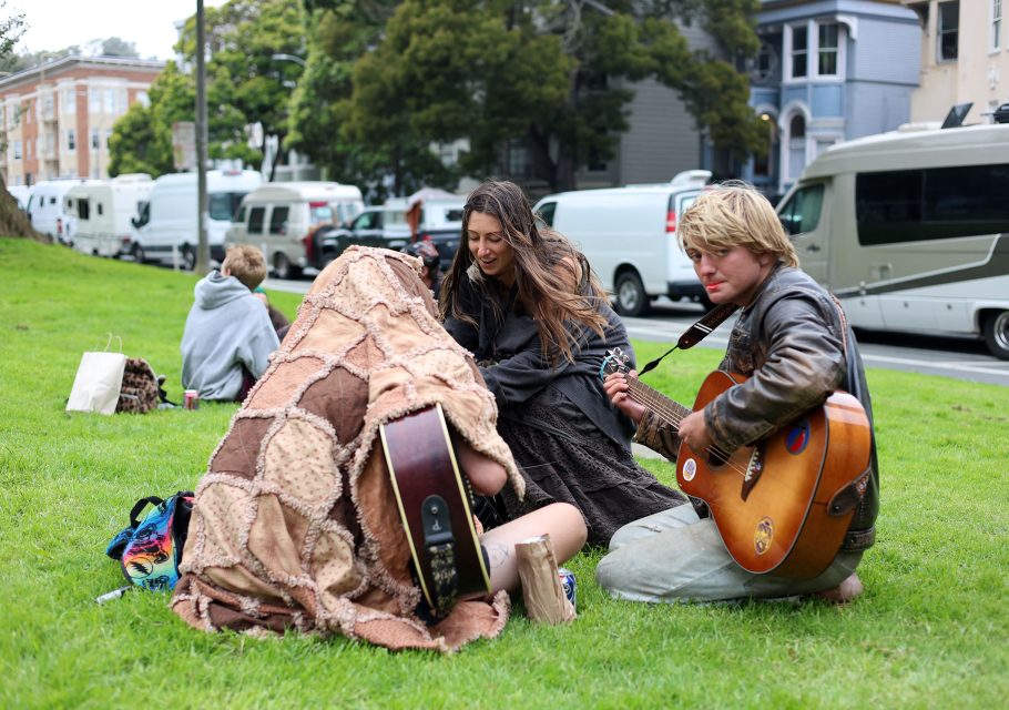 Three people sit on grass, two playing acoustic guitars and one covered with a patchwork blanket, with RVs and houses in the background.