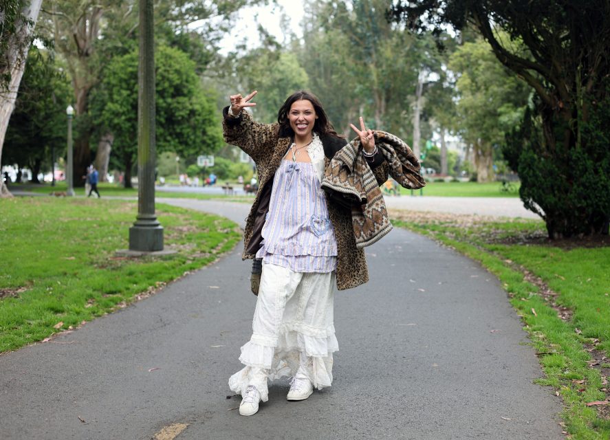 A woman walks on a park path, smiling and holding up peace signs with both hands. She is wearing a layered dress, a patterned coat, and white sneakers. Trees and grass surround her.