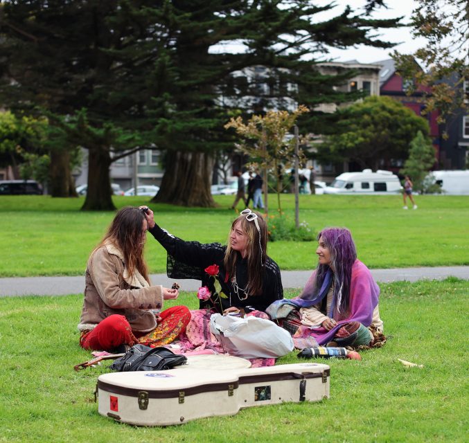 Three people sit on the grass in a park, talking and smiling, with a guitar case and bags in front of them. Trees and buildings are visible in the background.