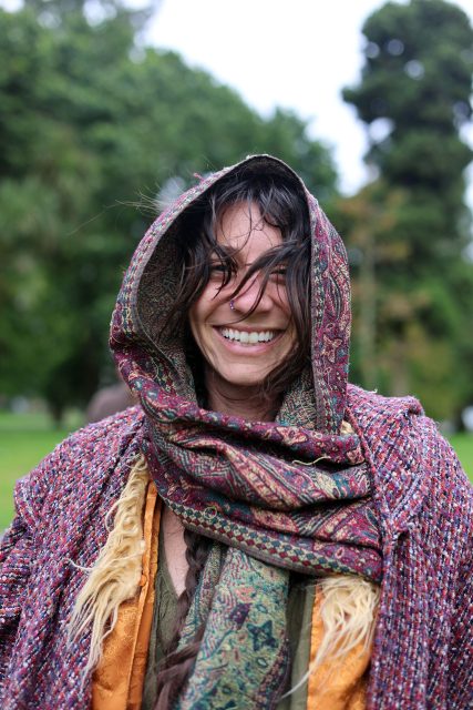 A woman standing outdoors, smiling, with windblown hair, wearing a patterned shawl and hood. Trees and greenery are visible in the blurred background.