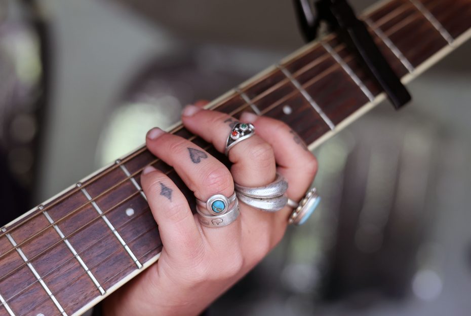 A close-up of a person's hand with rings and tattoos pressing guitar strings on the fretboard.