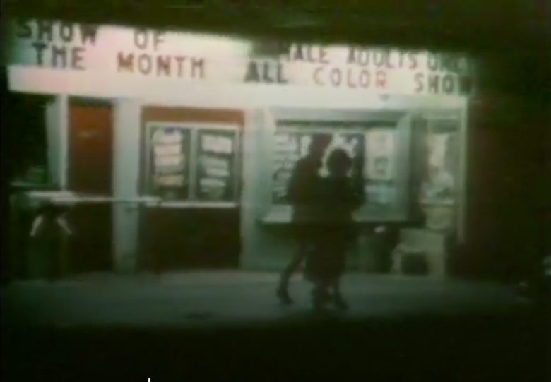 Two people walk in front of a theater at night with illuminated marquees reading "SHOW OF THE MONTH" and "ALL COLOR SHOW." Posters are displayed in glass cases by the entrance.
